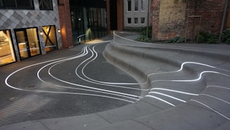 Curved concrete seating with embedded white LED lines creates a modern, artistic ambiance in an urban courtyard. The space is bordered by buildings with mixed architectural designs, including large windows and a checkerboard facade. Greenery and brick walls add contrast to the setting.