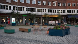 A public square featuring outdoor seating arrangements with tables and chairs, surrounded by large planters filled with colorful flowers. The backdrop includes a building with multiple windows and businesses, one of which is a 7-Eleven. The area is paved with stones, creating a structured, urban atmosphere.