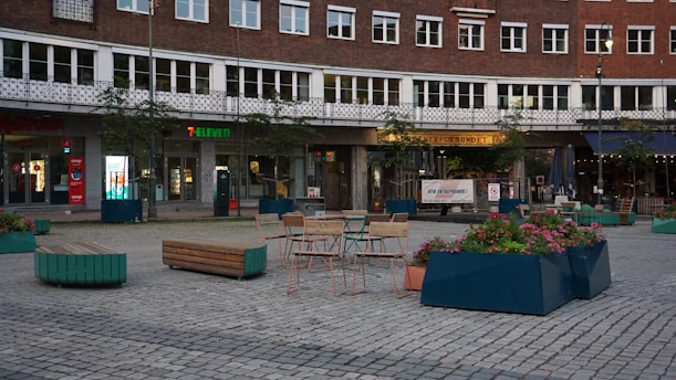 A public square featuring outdoor seating arrangements with tables and chairs, surrounded by large planters filled with colorful flowers. The backdrop includes a building with multiple windows and businesses, one of which is a 7-Eleven. The area is paved with stones, creating a structured, urban atmosphere.