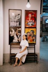 A woman in a white traditional dress sits on a wooden bench in a hallway. Above her are four movie posters, displaying various film scenes. A vintage style light bulb hangs from the ceiling, illuminating the scene.