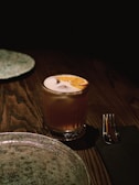 A cocktail in a glass with a slice of orange on top, set on a wooden table with dim lighting. Nearby, there is a ceramic plate and a fork placed on a black napkin.