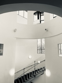 A modern, minimalist interior featuring a spiral staircase with metal railings. The walls are white, and natural light enters through tall, large windows, casting shadows on the smooth surfaces. Circular light fixtures and surveillance cameras are mounted on the walls. The architectural design emphasizes clean lines and open space.