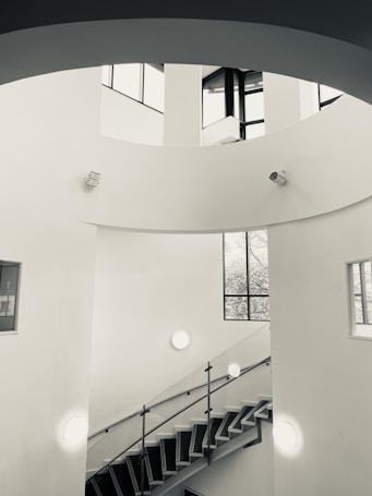 A modern, minimalist interior featuring a spiral staircase with metal railings. The walls are white, and natural light enters through tall, large windows, casting shadows on the smooth surfaces. Circular light fixtures and surveillance cameras are mounted on the walls. The architectural design emphasizes clean lines and open space.