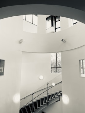A modern, minimalist interior featuring a spiral staircase with metal railings. The walls are white, and natural light enters through tall, large windows, casting shadows on the smooth surfaces. Circular light fixtures and surveillance cameras are mounted on the walls. The architectural design emphasizes clean lines and open space.