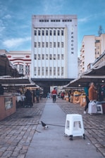 A tall, white building with vertical windows is in the background, with the word 'PROCON' displayed on its right side. The scene is set in an open-air market, with various stalls on either side of a stone-paved walkway. People are browsing the market, which appears to be selling fruits and vegetables. A pigeon and a white plastic stool are prominently seen in the foreground under a partly cloudy blue sky.