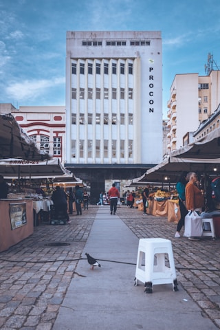 A tall, white building with vertical windows is in the background, with the word 'PROCON' displayed on its right side. The scene is set in an open-air market, with various stalls on either side of a stone-paved walkway. People are browsing the market, which appears to be selling fruits and vegetables. A pigeon and a white plastic stool are prominently seen in the foreground under a partly cloudy blue sky.