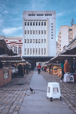 A tall, white building with vertical windows is in the background, with the word 'PROCON' displayed on its right side. The scene is set in an open-air market, with various stalls on either side of a stone-paved walkway. People are browsing the market, which appears to be selling fruits and vegetables. A pigeon and a white plastic stool are prominently seen in the foreground under a partly cloudy blue sky.