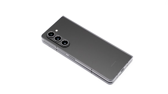 A modern smartphone with a sleek, foldable design is displayed against a white background. The device features three lenses and a flash in a vertical arrangement, highlighting its camera capabilities. Its minimalist and elegant appearance is emphasized by the smooth, dark surface.