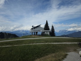 A modern building with the name 'Sonntagskogel' stands on a grassy hillside, surrounded by a scenic mountain range under a partly cloudy sky. Tall pine trees are situated next to the structure, and a pathway runs through the grass leading towards the horizon.