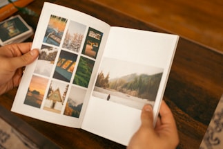 A person holding an open photo album featuring various photographs. The left page displays several small photos of landscapes and architectural structures organized in a grid. The right page shows a larger photo depicting a serene outdoor scene with trees and a river.