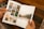 A person holding an open photo album featuring various photographs. The left page displays several small photos of landscapes and architectural structures organized in a grid. The right page shows a larger photo depicting a serene outdoor scene with trees and a river.