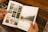 A person holding an open photo album featuring various photographs. The left page displays several small photos of landscapes and architectural structures organized in a grid. The right page shows a larger photo depicting a serene outdoor scene with trees and a river.
