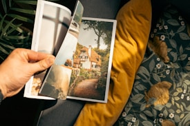 A hand holds open a magazine featuring a picturesque image of a cottage surrounded by lush greenery. The background includes a gray sofa with yellow and floral patterned cushions, creating a cozy setting.