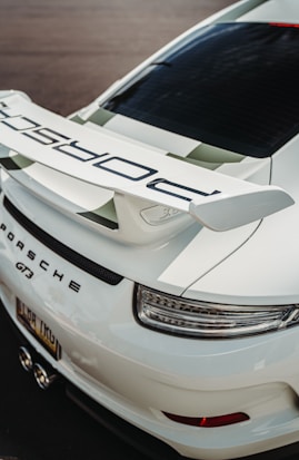 A close-up view of the rear section of a white sports car, featuring a prominent spoiler with sleek aerodynamic design. The car's branding is visible on the spoiler and the back of the vehicle, which includes large, sleek tail lights and dual exhaust pipes.