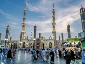 A grand mosque surrounded by towering minarets, with a large gathering of people at its entrance. The sky is partly cloudy, creating a peaceful atmosphere. Small vendor stalls line one side, adding a sense of everyday life and commerce to the scene.