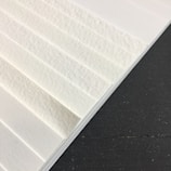 A close-up view of textured white paper sheets stacked at an angle against a dark surface. The surface of the paper has visible fibers, giving it a rough appearance.