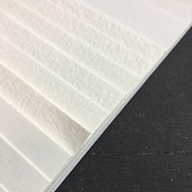 A close-up view of textured white paper sheets stacked at an angle against a dark surface. The surface of the paper has visible fibers, giving it a rough appearance.