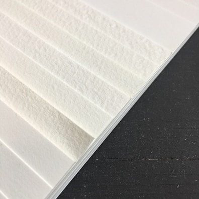 A close-up view of textured white paper sheets stacked at an angle against a dark surface. The surface of the paper has visible fibers, giving it a rough appearance.