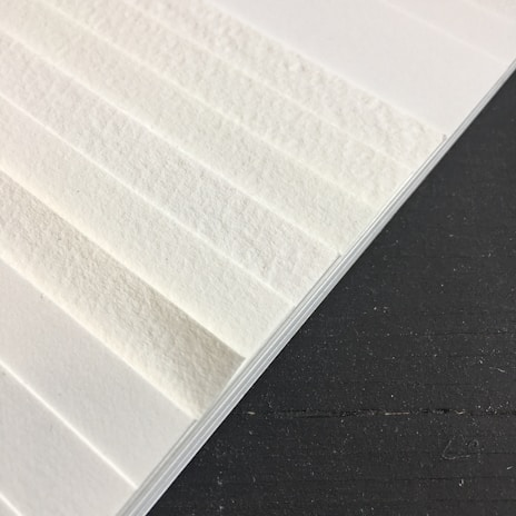A close-up view of textured white paper sheets stacked at an angle against a dark surface. The surface of the paper has visible fibers, giving it a rough appearance.