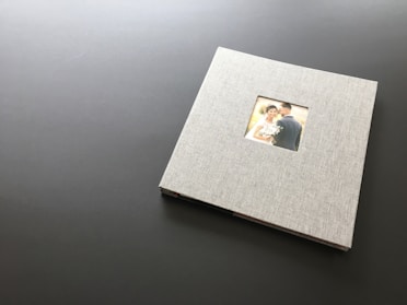 A closed photo album with a linen cover rests on a dark, flat surface. In the center of the album's cover is a small window displaying a photograph of a couple on their wedding day. The bride is holding a bouquet and smiling at the camera while the groom stands close beside her.