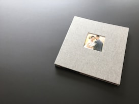A closed photo album with a linen cover rests on a dark, flat surface. In the center of the album's cover is a small window displaying a photograph of a couple on their wedding day. The bride is holding a bouquet and smiling at the camera while the groom stands close beside her.