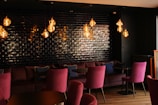 A stylish interior space featuring a set of plush magenta chairs and a comfortable couch along a wall tiled with dark, reflective bricks. Several pendant lights with warm, glowing bulbs hang from the ceiling, casting a cozy ambiance.