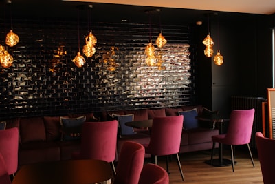 A stylish interior space featuring a set of plush magenta chairs and a comfortable couch along a wall tiled with dark, reflective bricks. Several pendant lights with warm, glowing bulbs hang from the ceiling, casting a cozy ambiance.