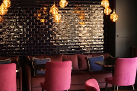 A cozy, modern lounge area featuring dark, glossy brick walls and vibrant, plush seating. The space is illuminated by several warm-toned pendant lights with artistic, patterned designs. Cushions in contrasting colors add a touch of elegance.