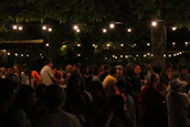 A lively community gathering outdoors with people sharing stories and music under string lights.