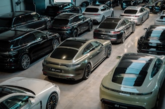 A showroom filled with multiple luxury cars parked in rows. The vehicles are sleek, modern, and exhibit a variety of models, predominantly in black, grey, and white finishes. The space is well-lit with reflections visible on the polished surfaces of the cars.