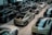 A showroom filled with multiple luxury cars parked in rows. The vehicles are sleek, modern, and exhibit a variety of models, predominantly in black, grey, and white finishes. The space is well-lit with reflections visible on the polished surfaces of the cars.