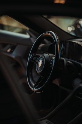 The image captures the interior of a luxury vehicle, focusing on the sleek black steering wheel with a logo at its center. The dashboard and other parts of the car's interior are visible, with modern and minimalist design elements highlighted by soft lighting.