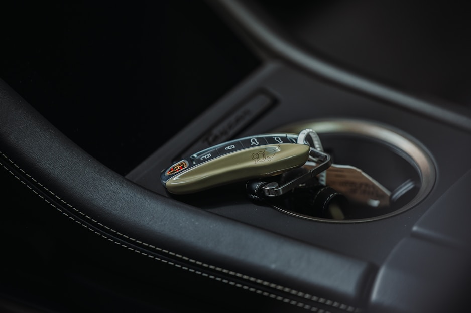 A luxurious car key fob rests in a car's cup holder, showing intricate design details and a logo. The setting includes sleek black leather with white stitching and a metallic interior accent.