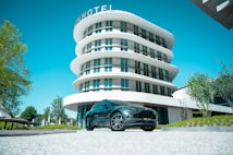 A modern architectural building with a curved design stands prominently in the background. In front of the building, a sleek black car is parked on a paved surface. The setting includes well-maintained greenery and a clear blue sky.
