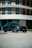 A sleek, dark-colored luxury car is parked in front of a modern building with large, vertically aligned windows and a curved architectural design. The car has a shiny finish, detailed with stylish wheels and distinctive headlights.