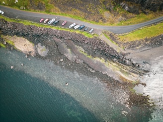 An aerial view of a coastal road with several vehicles parked along it. The road is lined by green grassy areas and rugged cliffs on one side and a rocky shoreline leading to a body of water on the other. The shoreline consists of dark rocks and patches of greenery.