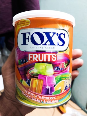 A hand holds a colorful tin of FOX's Crystal Clear Fruits which features vibrant imagery of fruits and multicolored candy. The tin is labeled with lemon, strawberry, blackcurrant, and orange flavors. The packaging includes bold blue and orange colors with images of fruit slices and candies.