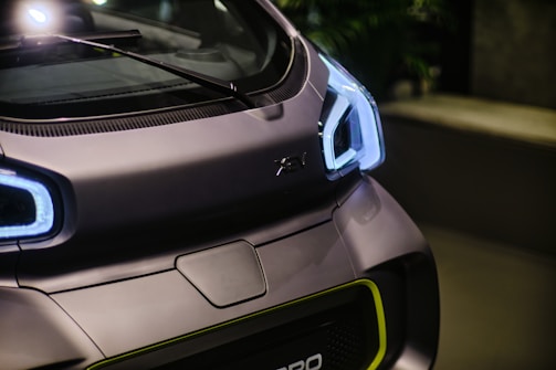 A close-up view of the front of a futuristic car with sleek, angular design. The vehicle features prominent LED headlights and a smooth matte finish. The windshield wiper is visible resting on the windshield.