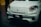 A close-up of the front of a white, modern electric vehicle with sleek design elements. The car features a prominent logo on its hood and uniquely shaped headlights. The license plate displays the text 'T03'.