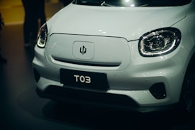 A close-up of the front of a white, modern electric vehicle with sleek design elements. The car features a prominent logo on its hood and uniquely shaped headlights. The license plate displays the text 'T03'.