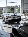 The image features a showroom with large windows allowing natural light to illuminate a sleek white sports car. Several other vehicles are also present, reflecting a modern and luxurious setting. The outside view shows office buildings and greenery.