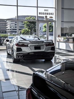 The image features a showroom with large windows allowing natural light to illuminate a sleek white sports car. Several other vehicles are also present, reflecting a modern and luxurious setting. The outside view shows office buildings and greenery.
