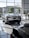 The image features a showroom with large windows allowing natural light to illuminate a sleek white sports car. Several other vehicles are also present, reflecting a modern and luxurious setting. The outside view shows office buildings and greenery.