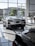 The image features a showroom with large windows allowing natural light to illuminate a sleek white sports car. Several other vehicles are also present, reflecting a modern and luxurious setting. The outside view shows office buildings and greenery.