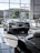The image features a showroom with large windows allowing natural light to illuminate a sleek white sports car. Several other vehicles are also present, reflecting a modern and luxurious setting. The outside view shows office buildings and greenery.