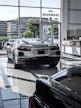 The image features a showroom with large windows allowing natural light to illuminate a sleek white sports car. Several other vehicles are also present, reflecting a modern and luxurious setting. The outside view shows office buildings and greenery.