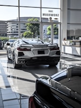 The image features a showroom with large windows allowing natural light to illuminate a sleek white sports car. Several other vehicles are also present, reflecting a modern and luxurious setting. The outside view shows office buildings and greenery.