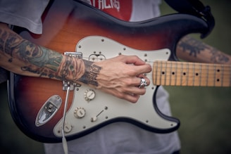 A person with tattooed arms is playing an electric guitar. The guitar has a sunburst finish and features multiple control knobs. The person is wearing a silver skull ring and the background appears to be outdoors, possibly in a park.