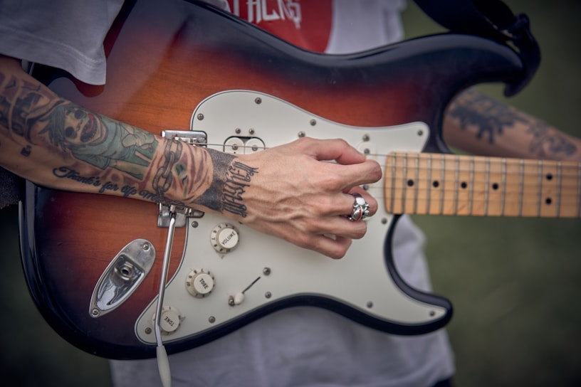 A person with tattooed arms is playing an electric guitar. The guitar has a sunburst finish and features multiple control knobs. The person is wearing a silver skull ring and the background appears to be outdoors, possibly in a park.