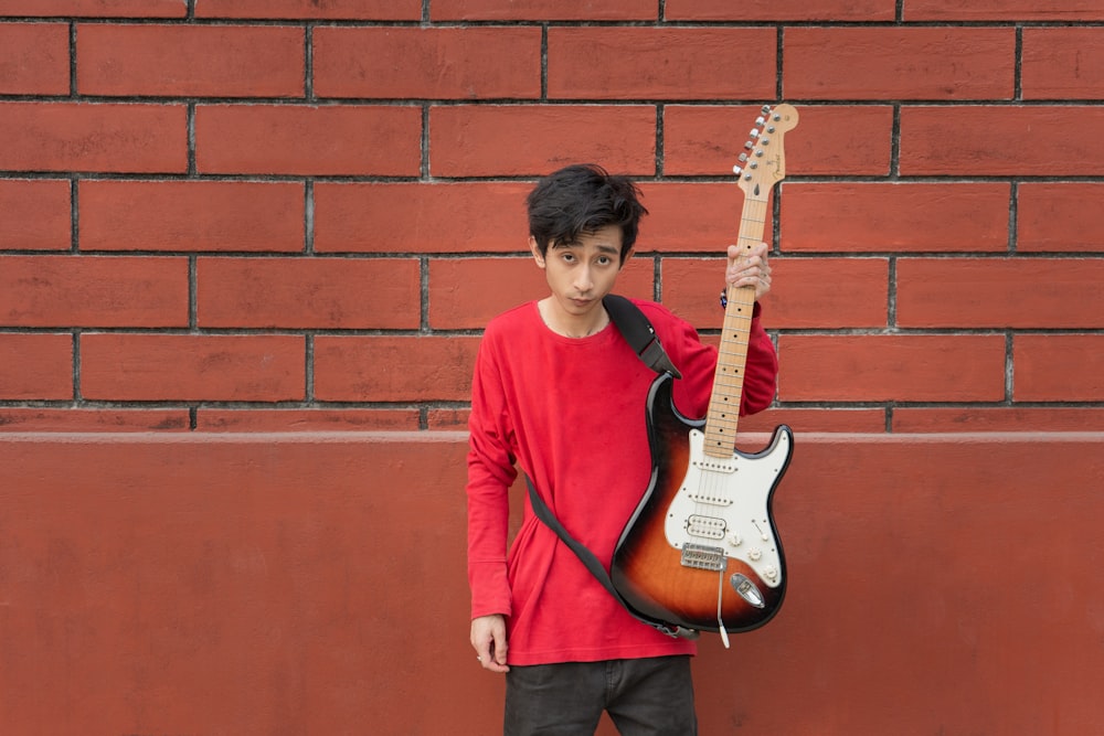 a young boy holding a guitar in front of a brick wall